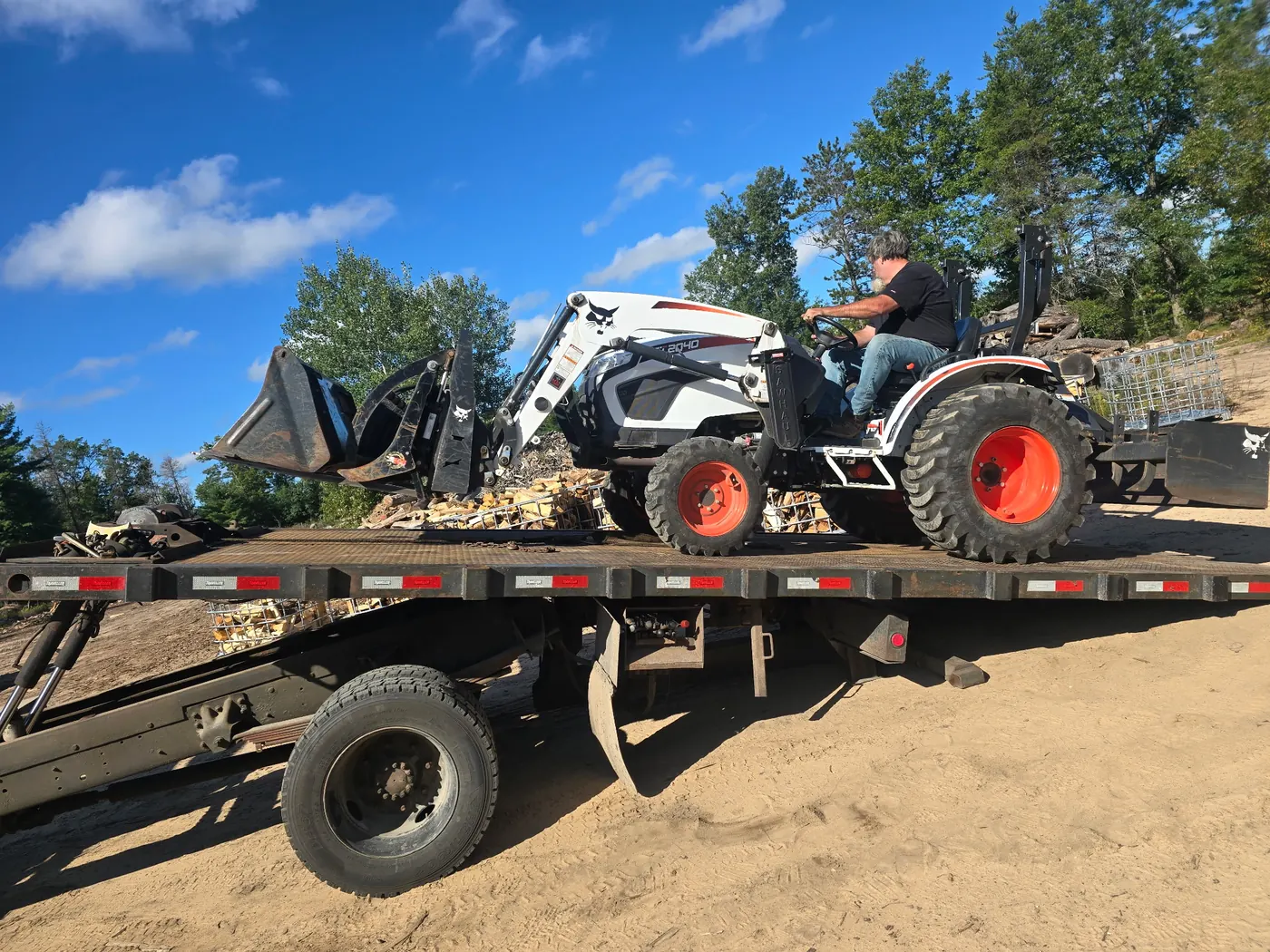 Bobcat compact tractor with front loader driving up onto a C&C Inc rollback flatbed tow truck in Northern Michigan