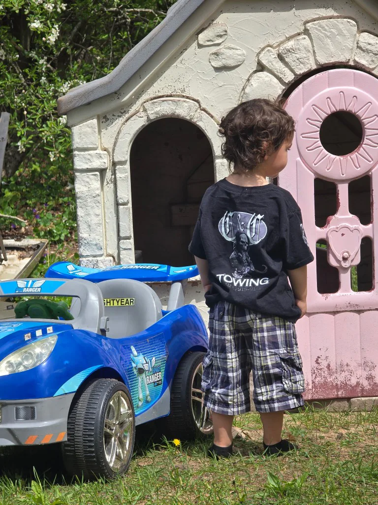 Young child in a C&C Towing shirt playing in the backyard with a toy car and playhouse